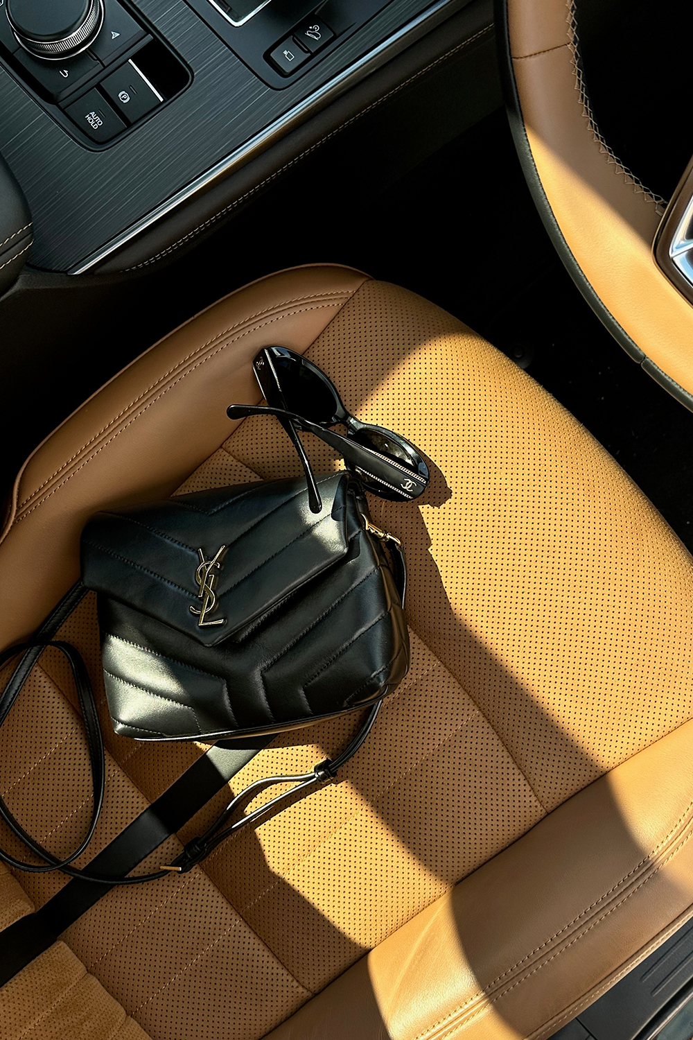 Black designer handbag and sunglasses resting on a tan leather car seat in warm natural light, creating a refined, minimalist luxury aesthetic.